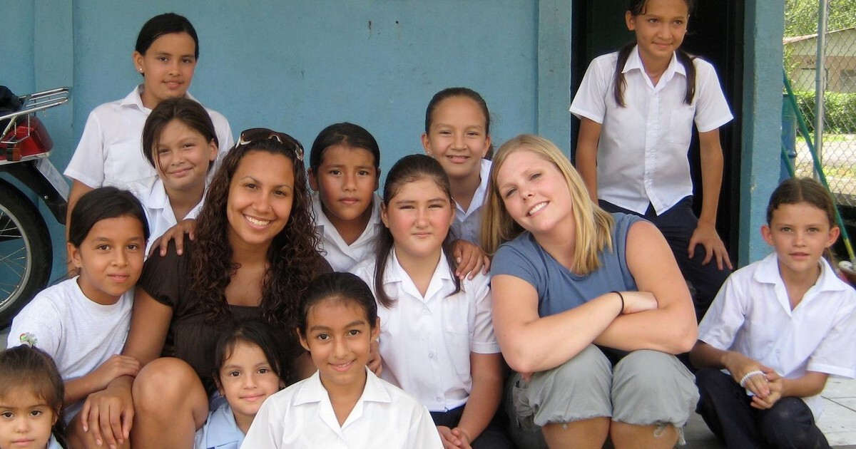 A volunteer teacher poses with a group of smiling schoolgirls in uniform outside a classroom building.