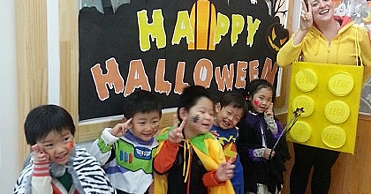 A smiling teacher in a homemade yellow building-block costume poses with five young students in Halloween costumes and face paint beside a hand-painted Happy Halloween classroom sign.