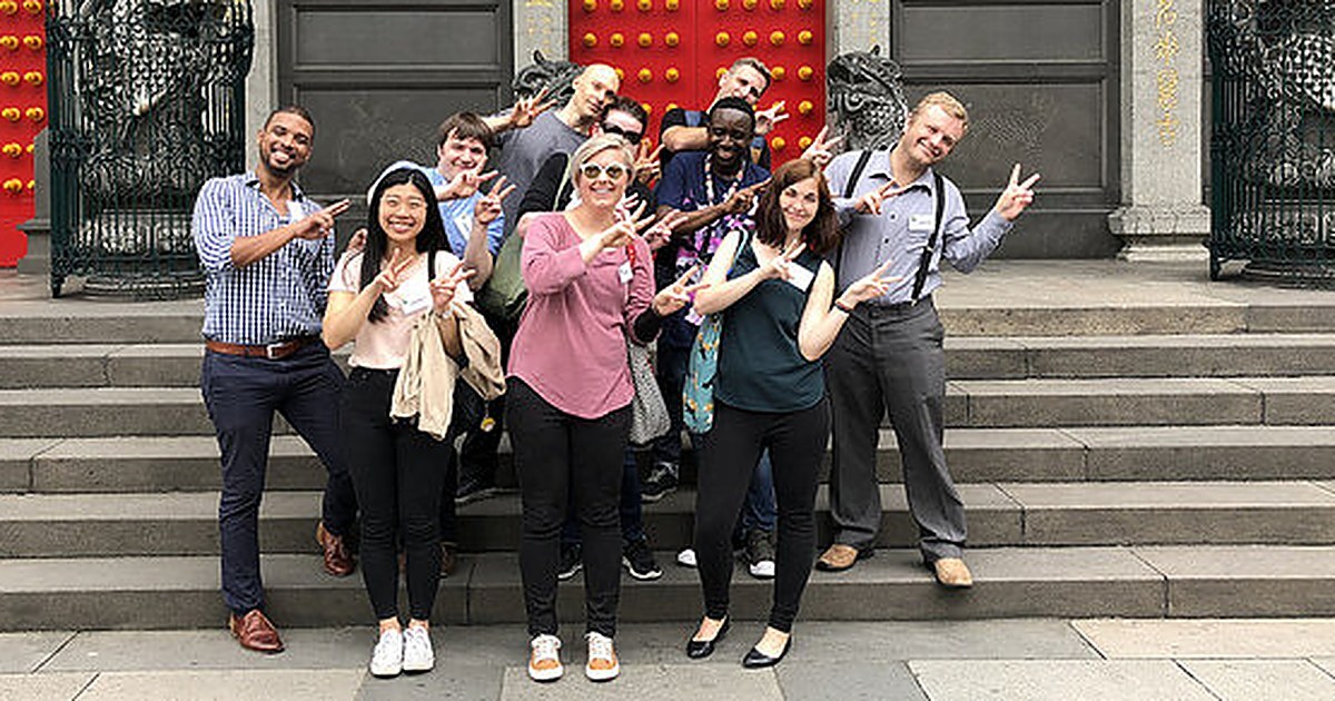 A diverse group of men and women teaching English abroad, posing together on the steps of a traditional red-doored temple.