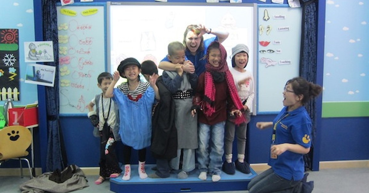 An English teacher laughing with a group of young students in costumes on a small classroom stage, with a teaching assistant joining in.