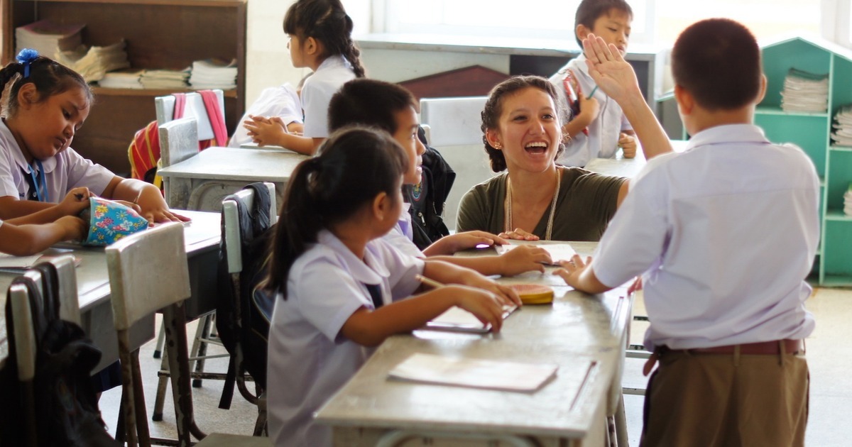 A smiling teacher sits with a group of young students at classroom desks as one boy raises his hand for a high five.